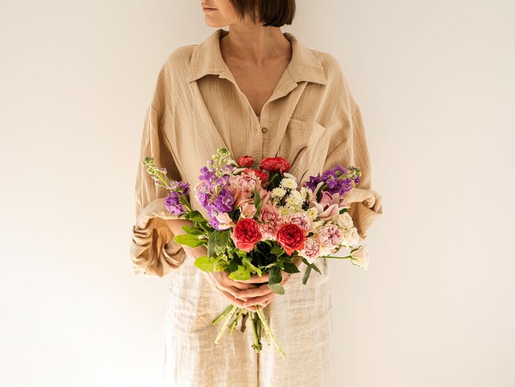 Schwitzen Schweiß Hitze Sommer Stoffe Frau mit kurzem braunem Haar hält einen bunten Strauß Blumen vor cremefarbenem Hintergrund und blickt zur Seite.