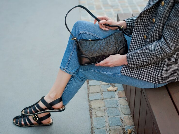 Schwarze Sommersandalen mit goldener Schnalle an schlanken Beinen in Jeans, Frau hält elegante Handtasche auf dem Schoß.