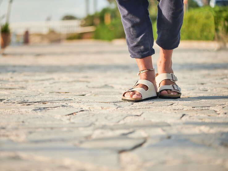 Sandalen Birkenstock Sommer Füße in beigen Birkenstock-Sandalen und hochgekrempelter Hose auf heller Natursteinpromenade bei warmem Abendlicht.