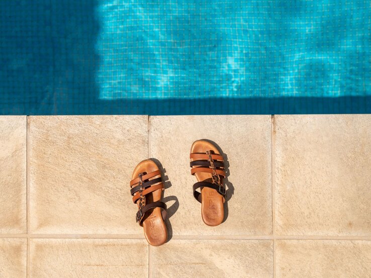 Ein Paar braune Sandalen liegt nebeneinander im Mallorca-Urlaub auf beigem Fliesenrand vor glitzernd blauem Pool im Sonnenlicht.