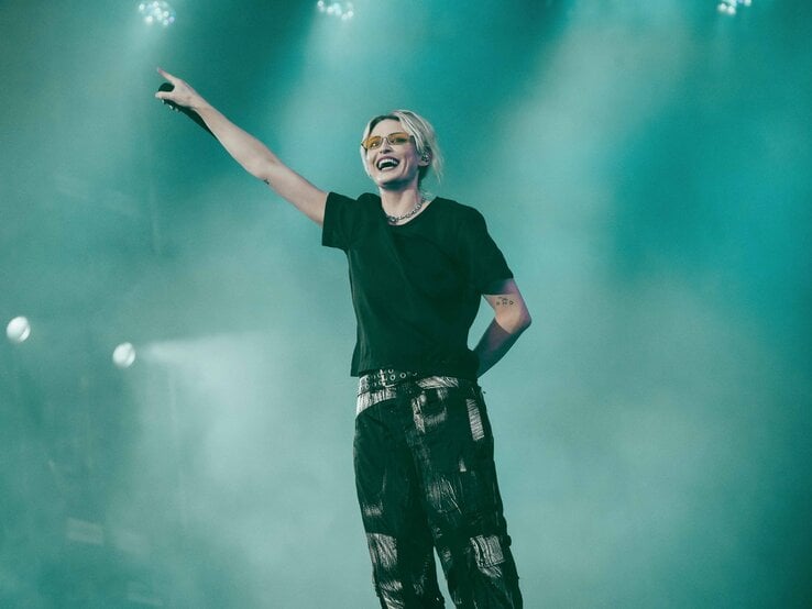 Linkin-Park-Sängerin Emily Armstrong mit Sonnenbrille und dunklem Outfit zeigt lachend auf die jubelnde Menge in türkisfarbenem Bühnenlicht.