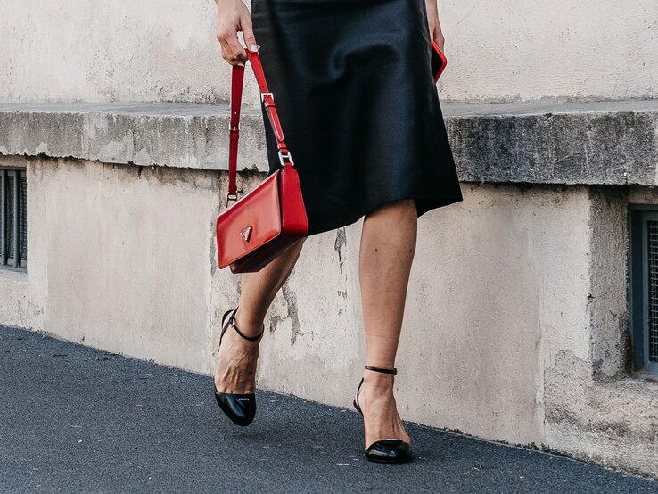 Frau in schwarzem Midirock geht auf Gehweg, trägt rote Handtasche und schwarze Sandalen mit Riemchen.