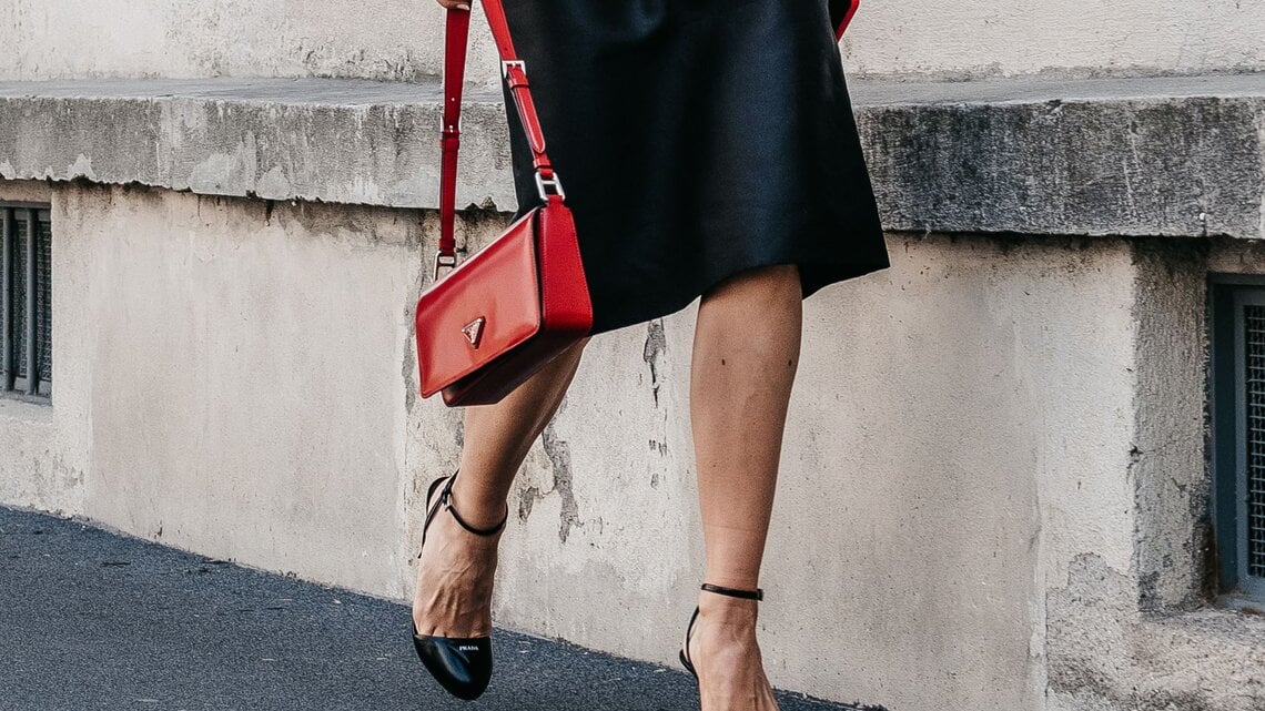 Frau in schwarzem Midirock geht auf Gehweg, trägt rote Handtasche und schwarze Sandalen mit Riemchen.