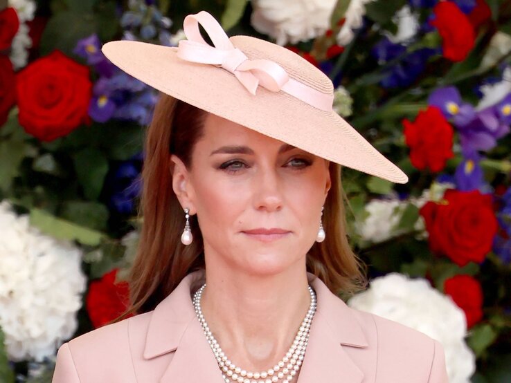 Elegant gekleidete Prinzessin Kate mit Perlenkette und rosafarbenem Hut blickt ernst vor einem Hintergrund aus roten und weißen Blumen.