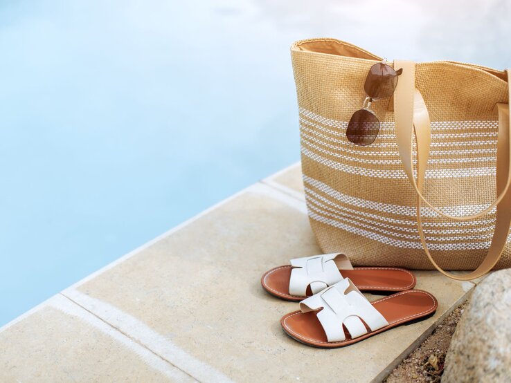 Sommerliche Szene mit gestreifter Basttasche, brauner Sonnenbrille und weißen Ledersandalen neben klarem Poolwasser.