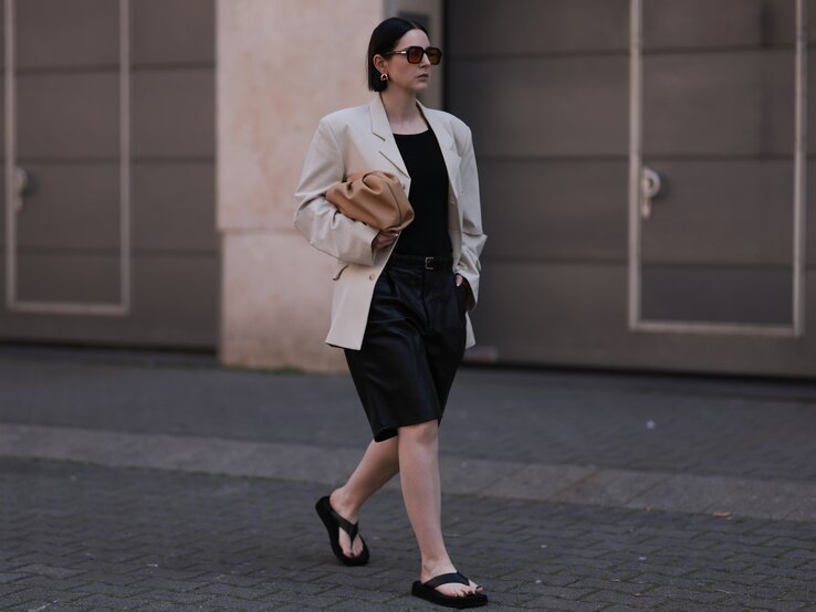 Zu sehen ist eine Frau mit einem Bob, die schwarze Zehensandalen, eine Bermudashorts, ein schwarzes Top und einen Cremfarbenen Blazer.