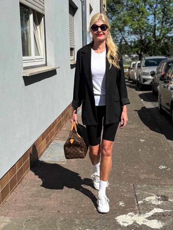 Renate Zott in schwarzem Blazer und Radlerhose geht mit Designerhandtasche und Sonnenbrille an Hausfassade entlang. | © Renate Zott 