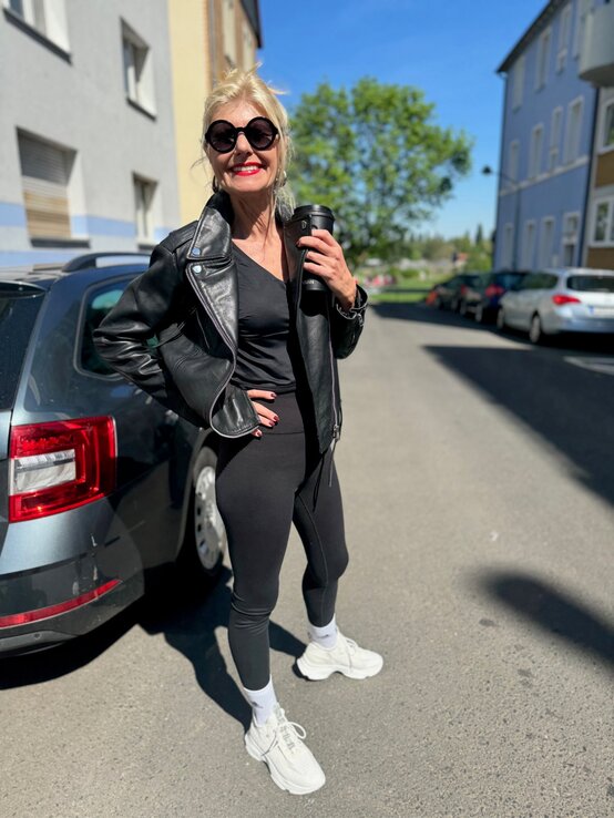 Renate Zott in schwarzer Lederjacke, Leggings und weißen Sneakern steht mit Coffee-to-go auf sonniger Straße. | © Renate Zott 
