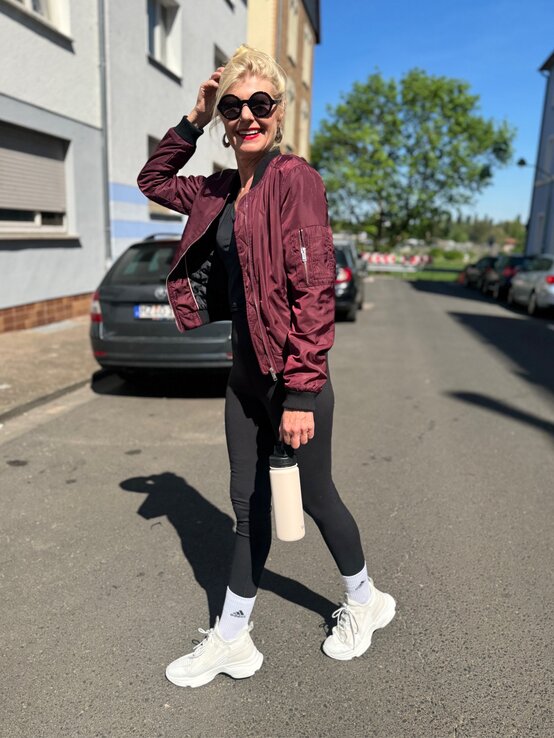 Renate Zott in bordeauxfarbener Bomberjacke und schwarzer Leggings spaziert lachend mit Trinkflasche durch sonnige Straße. | © Renate Zott 