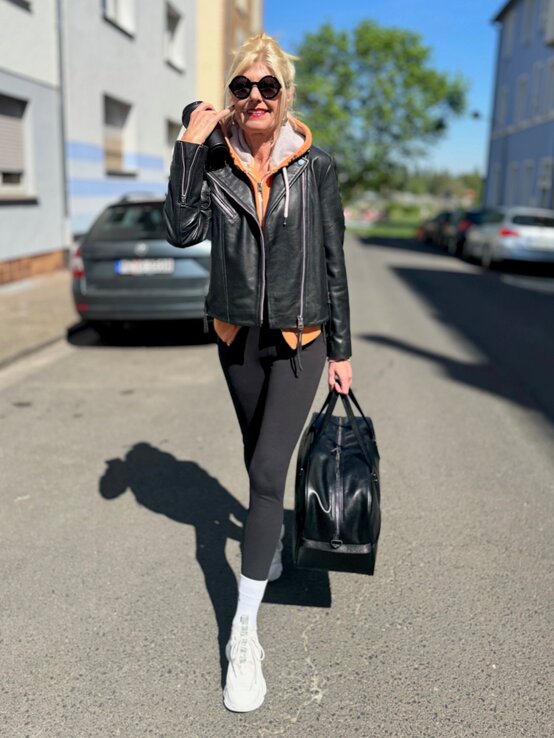 Renate Zott in schwarzer Lederjacke, Leggings und weißen Sneakern geht mit Sporttasche und Coffee-to-go über sonnige Straße. | © Renate Zott 