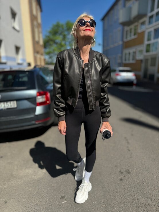 Renate Zott in schwarzer Pilotenjacke, Leggings und weißen Sneakers blickt lächelnd mit Sonnenbrille in den strahlend blauen Himmel. | © Renate Zott 