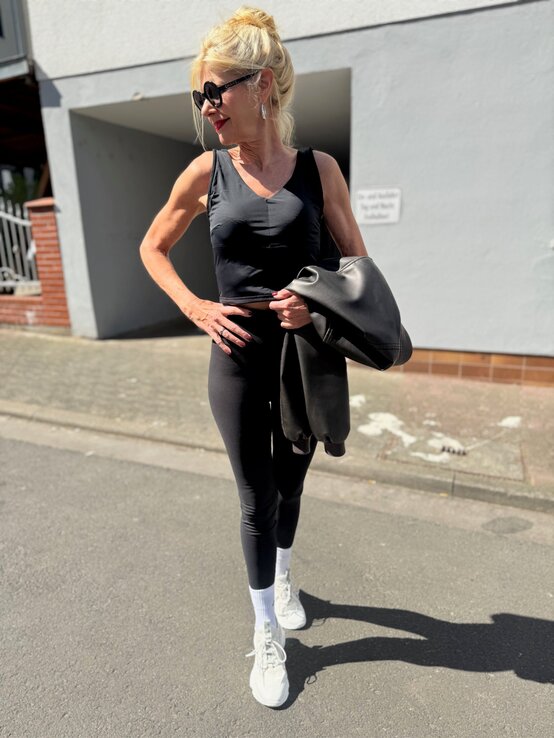 Renate Zott mit Dutt und Sonnenbrille steht in schwarzem Sportoutfit und weißen Sneakers selbstbewusst im Sonnenlicht. | © Renate Zott 