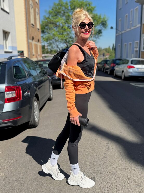 Renate Zott mit Sonnenbrille, orangefarbener Jacke und schwarzem Sportoutfit läuft lachend mit Rucksack durch Wohnstraße. | © Renate Zott 