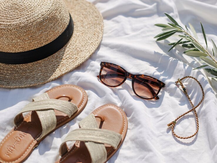 Strohhut, Sandalen, Sonnenbrille und ein goldenes Armband liegen sommerlich arrangiert auf weißem Tuch im Sonnenlicht.