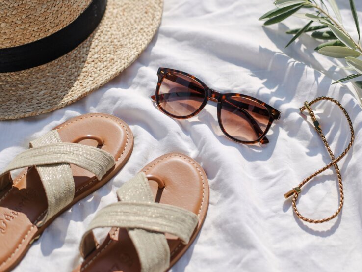 Strohhut, Sandalen, Sonnenbrille und ein goldenes Armband liegen sommerlich arrangiert auf weißem Tuch im Sonnenlicht.