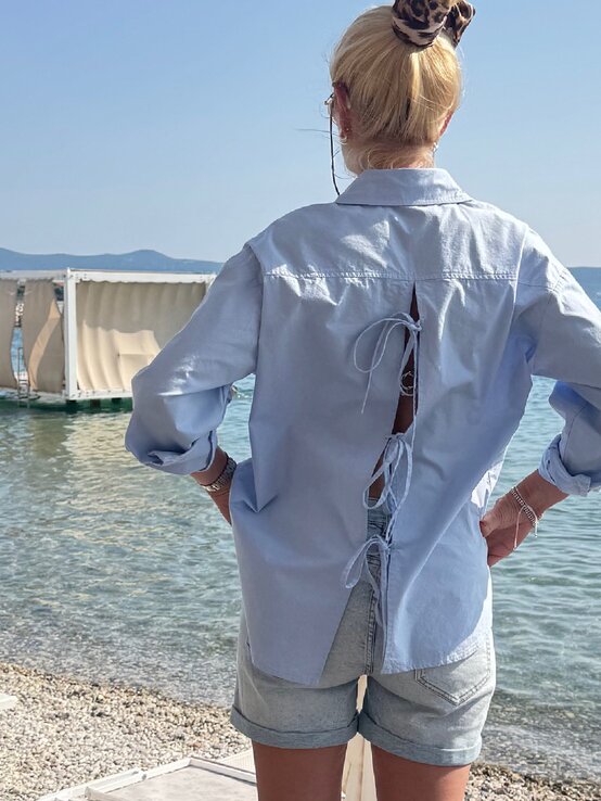 Frau in lässigem Sommeroutfit steht mit den Händen in den Hüften am Strand, während Menschen im türkisfarbenen Meer planschen. | © Renate Zott