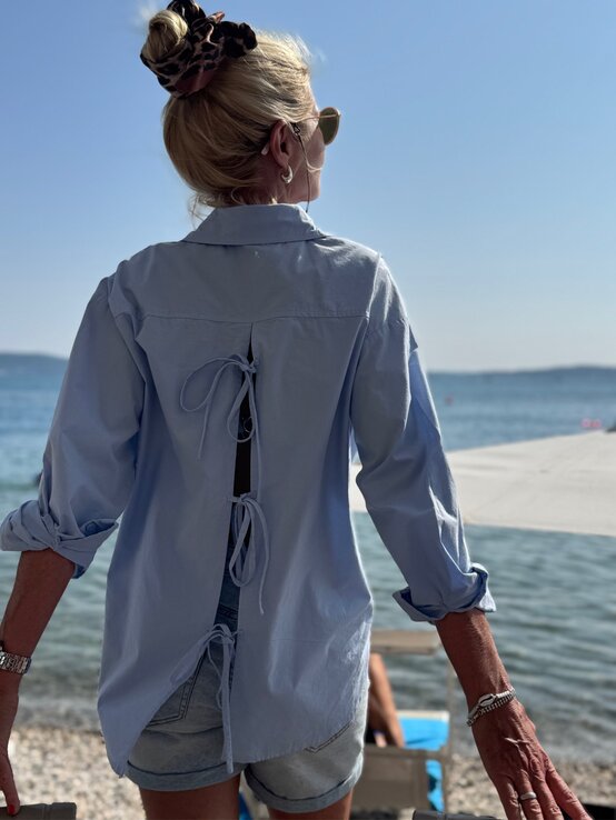 Blonde Frau mit Sonnenbrille und Leoparden-Haargummi steht am Kiesstrand, trägt ein locker gebundenes blaues Hemd und blickt aufs Meer. | © Renate Zott