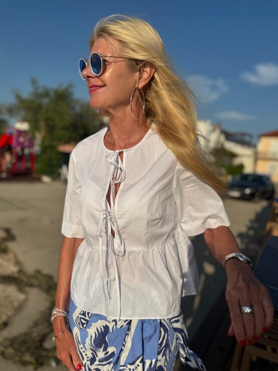 Frau mit langen blonden Haaren trägt eine weiße Bluse mit Kordelverschluss und blau-weiß gemusterten Rock in sommerlicher Abendstimmung. | © Renate Zott