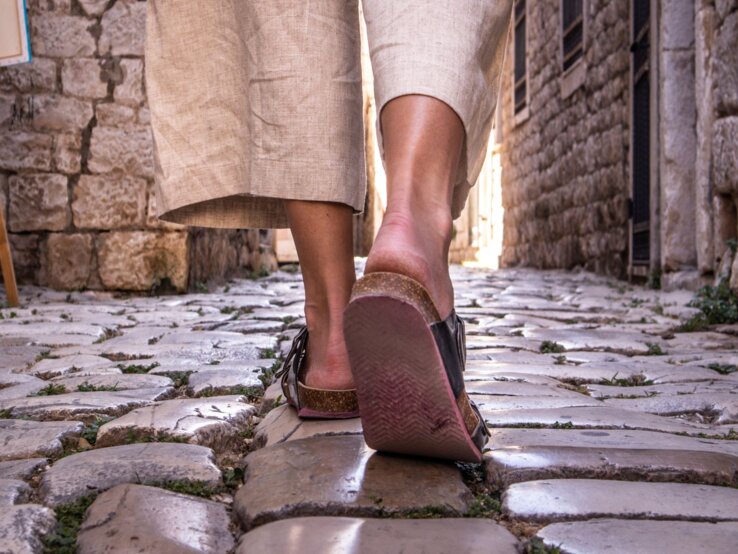 Nahaufnahme von Beinen in beigen Leinenhosen und Sandalen, die über glänzende Kopfsteinpflaster in einer engen Altstadtgasse laufen.