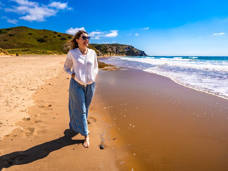 Portugal Hose Tchibo Sommer Eine Frau in weißer Bluse und blauer Hose spaziert barfuß am sonnigen Sandstrand entlang, während kleine Wellen ans Ufer schlagen.
