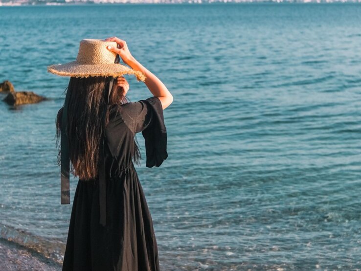Eine Frau mit langem dunklem Haar und Strohhut steht in einem schwarzen Kleid am ruhigen Meer, dahinter Berge und eine Küstenstadt in Griechenland.