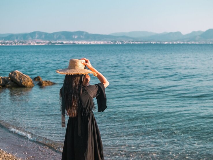 Mode wie in Griechenland Eine Frau mit langem dunklem Haar und Strohhut steht in einem schwarzen Kleid am ruhigen Meer, dahinter Berge und eine Küstenstadt in Griechenland.