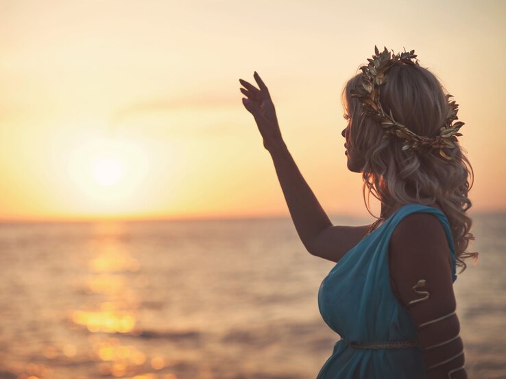 Eine junge Frau mit blonden Locken, goldener Lorbeerkrone und blauem Kleid hebt die Hand vor einem goldenen Sonnenuntergang am Meer.