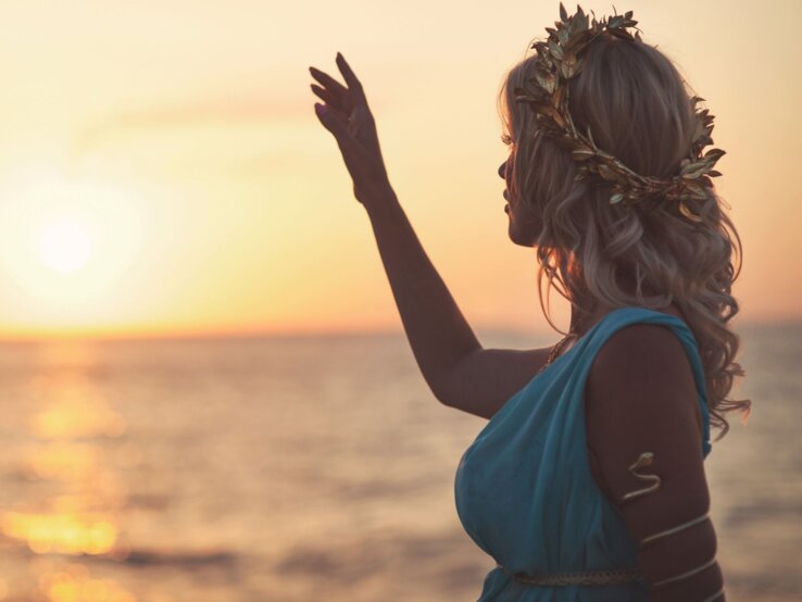 Eine junge Frau mit blonden Locken, goldener Lorbeerkrone und blauem Kleid hebt die Hand vor einem goldenen Sonnenuntergang am Meer.