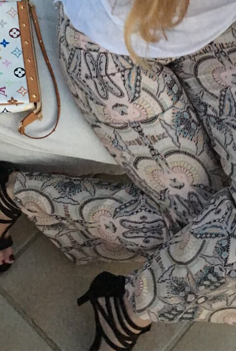 Eine Frau in bedruckter Hose, schwarzen High Heels und weißem Shirt hält ein türkisfarbenes Getränk, daneben eine Designer-Handtasche.