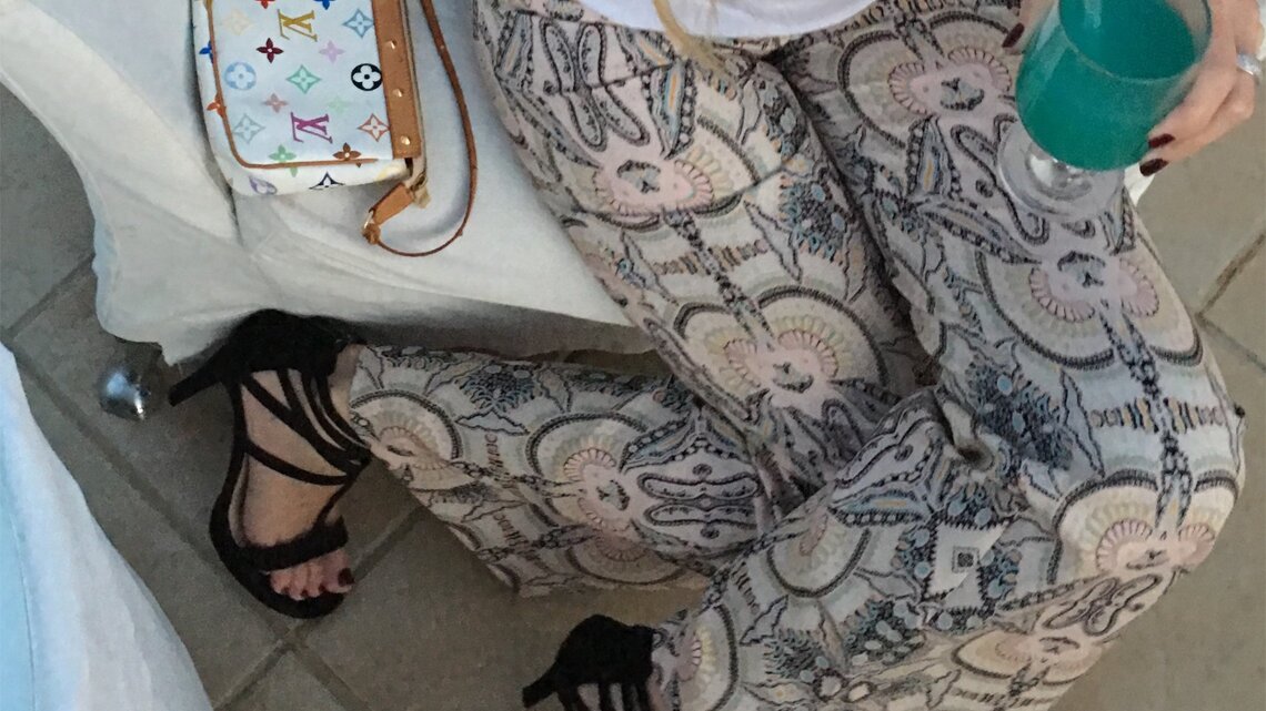 Eine Frau in bedruckter Hose, schwarzen High Heels und weißem Shirt hält ein türkisfarbenes Getränk, daneben eine Designer-Handtasche.