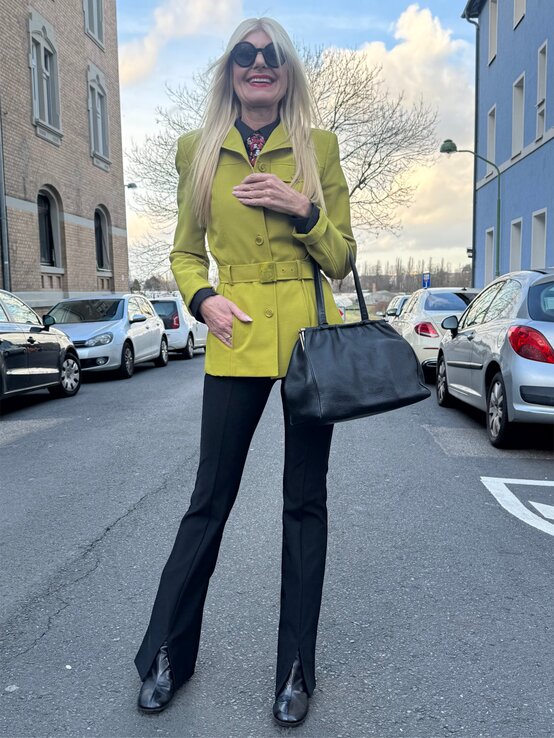 Eine Frau mit langen blonden Haaren trägt einen limettengrünen Mantel, schwarze Hose, Stiefeletten und eine schwarze Handtasche auf der Straße. | © Renate Zott