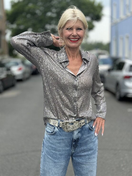 Eine Frau in glitzernder silberner Bluse mit Verzierungen, heller Jeans und auffälligem Gürtel posiert lässig auf einer Stadtstraße. | © Renate Zott