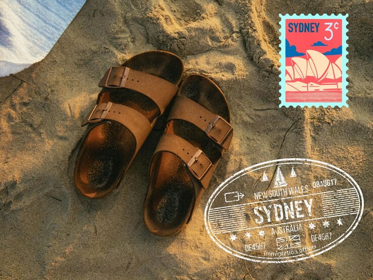 Paar braune Sandalen liegt auf sonnigem Sand neben einem Sydney-Briefmarkenmotiv und einem Reise-Stempel-Design.