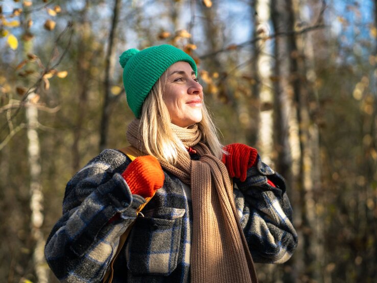 Eine Person mit blonden Haaren trägt eine grüne Mütze, rote Handschuhe, einen braunen Schal und eine karierte Jacke in einem herbstlichen Birkenwald.