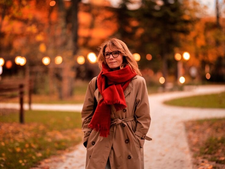 Scandinavian Scarf Trend Schal Herbst Eine Person mit offenem, mittellangem Haar trägt einen beigefarbenen Mantel mit rotem Schal auf einem herbstlichen Parkweg bei Dämmerung.