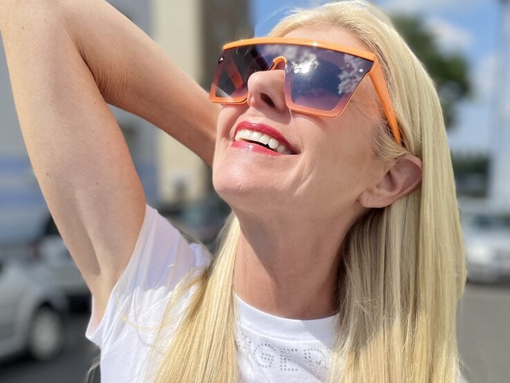 Nahaufnahme von langem platinblondem Haar, weißem T-Shirt und orange gefärbten Sonnenbrillenbügeln vor sommerlicher Kulisse. | © Renate Zott