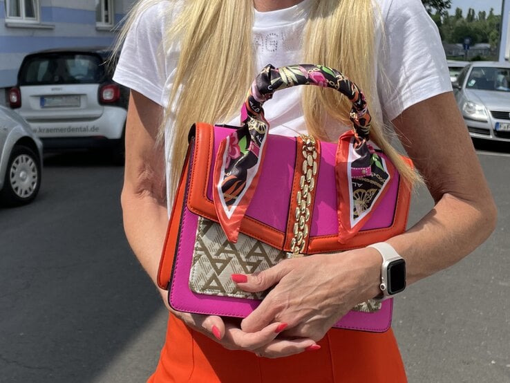 Elegante Handtasche in kräftigem Pink und Orange mit goldenen Ketten und dekorativen Tüchern, gehalten zu einem klassischen weißen T-Shirt. | © Renate Zott