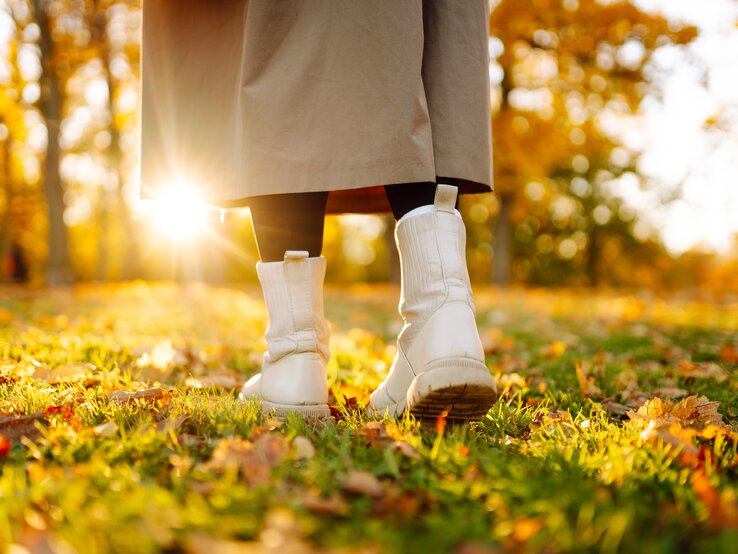 Weiße Stiefel einer Person in schwarzer Strumpfhose und beigem Mantel spazieren bei goldener Herbstsonne über eine Wiese mit Laub.
