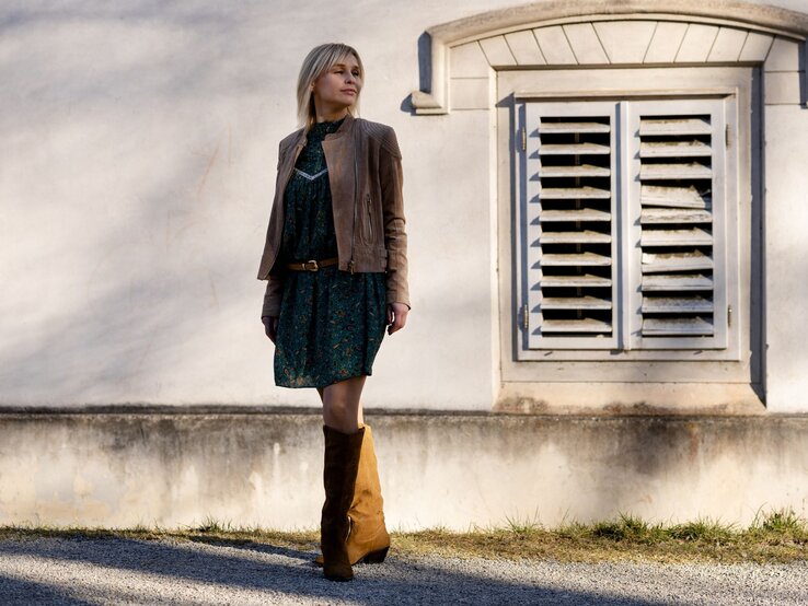 Frau mit langen blonden Haaren trägt braune Wildlederstiefel, grünes gemustertes Kleid und braune Lederjacke vor sonniger Hausfassade.