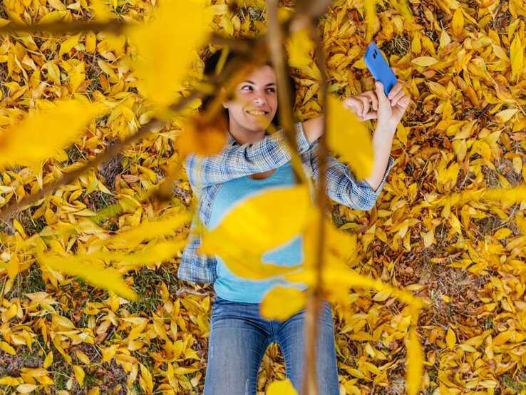 Pollunder von Tschibo Herbst Eine Frau liegt im bunten Laub auf dem Boden und trägt ein blaues Oberteil und lächelt.