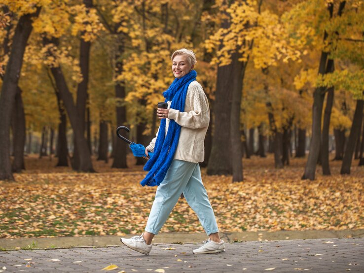Eine Person im cremefarbenen Pullover und blauer Jeans spaziert mit Regenschirm und Kaffee durch einen herbstlichen Park voller Laub.