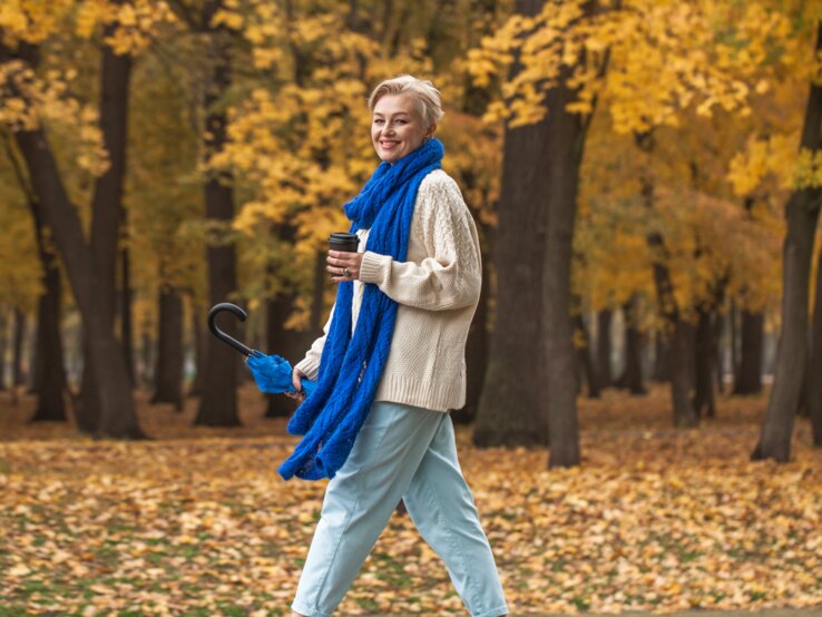 Eine Person im cremefarbenen Pullover und blauer Jeans spaziert mit Regenschirm und Kaffee durch einen herbstlichen Park voller Laub.