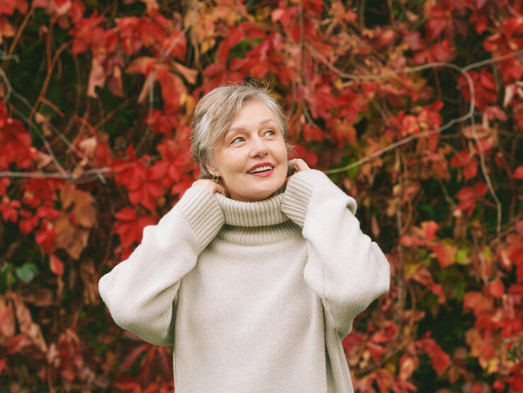 Mode To-dos Herbst Eine Person in cremeweißem Rollkragenpullover steht vor einer herbstlichen Wand aus roten und grünen Blättern.