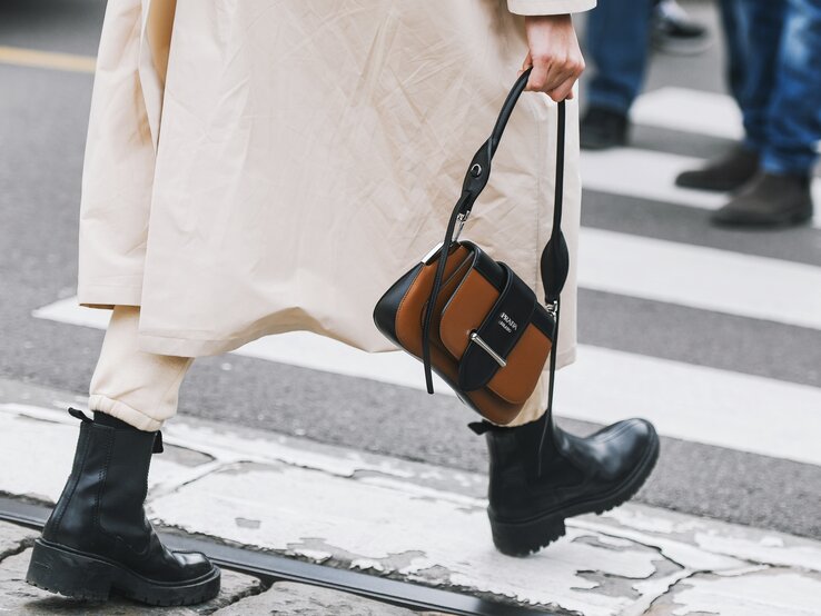 Schwarze klobige Stiefel und beige Hose unter langem Mantel, Person hält eine braun-schwarze Designerhandtasche, Gehweg im Hintergrund.