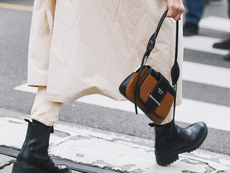 Schwarze klobige Stiefel und beige Hose unter langem Mantel, Person hält eine braun-schwarze Designerhandtasche, Gehweg im Hintergrund.