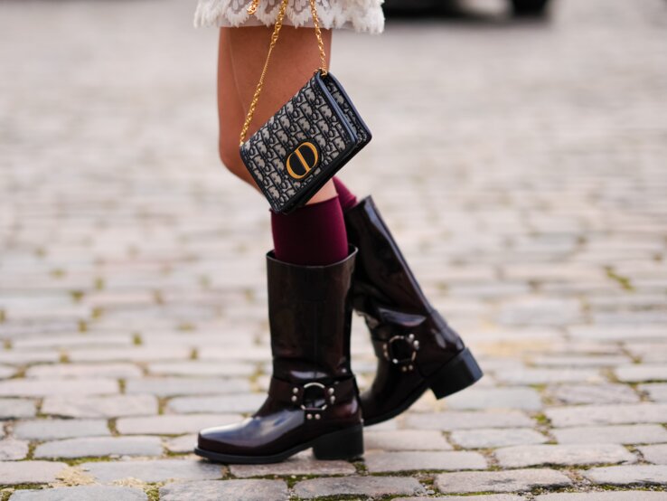 Flache Stiefel Trend Herbst 2025 Bonprix Detailaufnahme von braunen Lackstiefeln, bordeauxroten Strümpfen und einer schwarz-gemusterten Handtasche mit goldener Kette auf steinigem Untergrund.