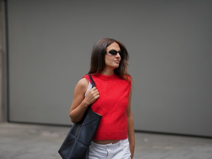Pullunder Trend Herbst Frau in auffälligem rotem Pullover, weißer Hose und schwarzer Tragetasche steht seitlich auf grauem Steinboden vor einer Wand.
