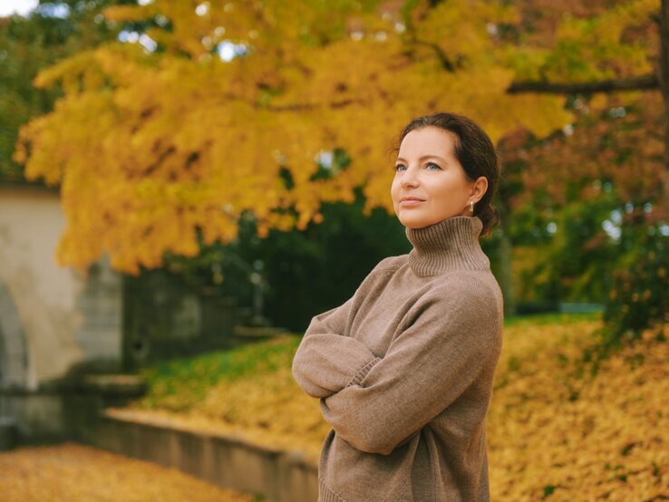 Rollkragen Pullover stylen Eine Person in einem hellbraunen Rollkragenpullover steht entspannt vor Herbstbäumen mit goldgelbem Laub und einem steinernen Torbogen.