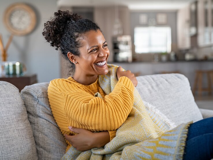 Pullover Herbst Person mit lockigem Haar in gelbem Strickpullover sitzt auf grauem Sofa, in eine Decke gewickelt, im hellen Wohnzimmer.