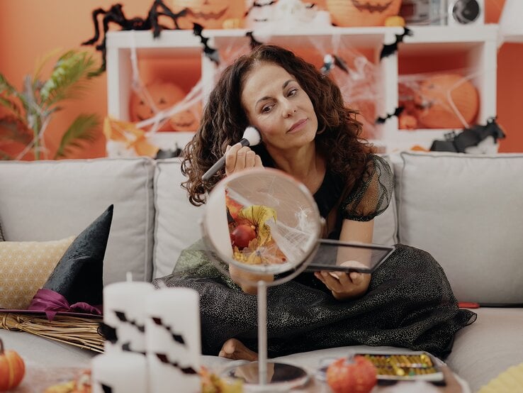 Last Minute Halloween Kostüm DIY Eine Frau in glitzerndem schwarzen Kleid sitzt auf einem grauen Sofa, mit Halloween-Dekoration und Kürbissen im orangefarbenen Hintergrund.